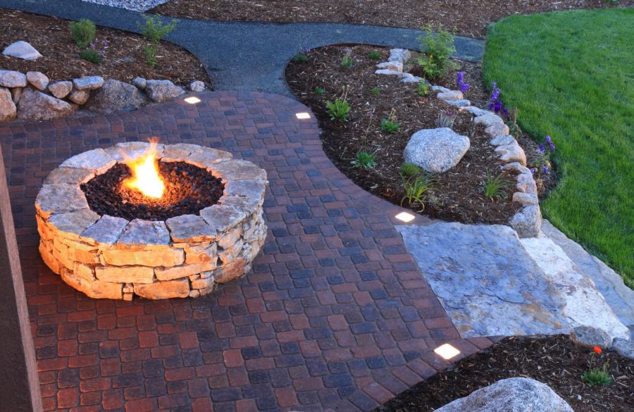 Stone fire pit burning on a brick patio in a landscaped backyard garden